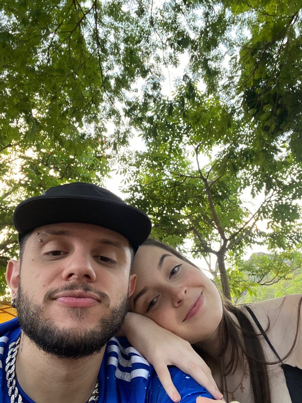 Selfie do casal sob árvores, Marta com a cabeça apoiada no ombro de Matheus