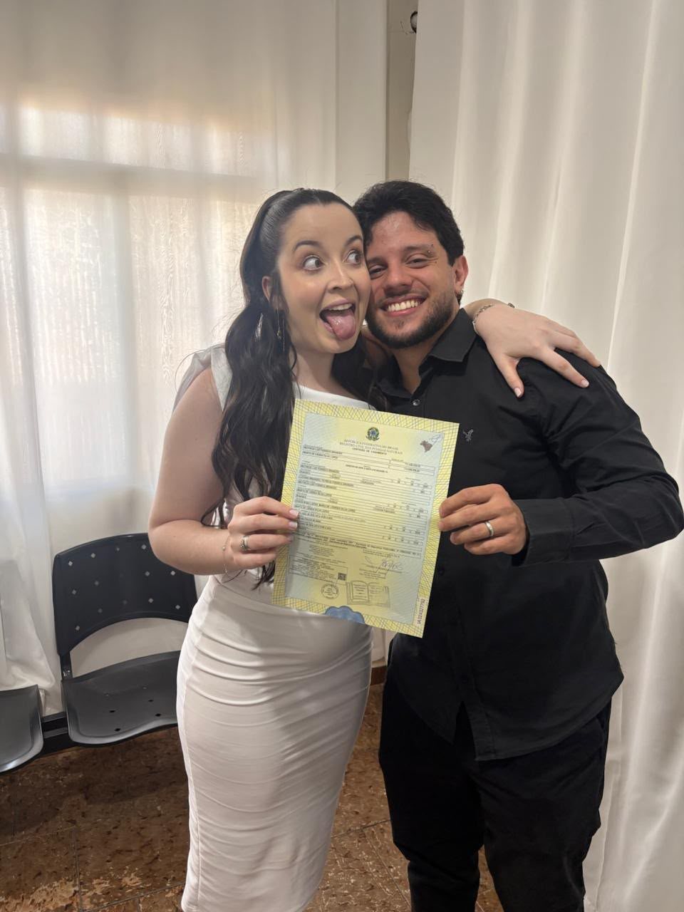 Marta e Matheus abraçados segurando a certidão do casamento civil, ela de vestido branco fazendo careta