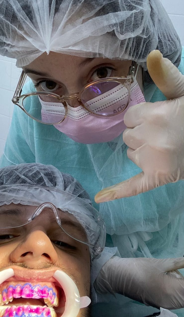 Marta de touca e máscara no consultório fazendo sinal de positivo, com Matheus deitado na cadeira odontológica durante clareamento