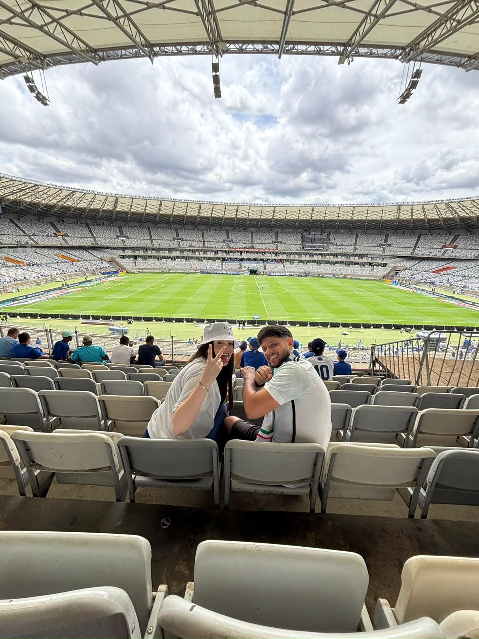 Marta e Matheus sentados na arquibancada do estádio Mineirão com o gramado ao fundo